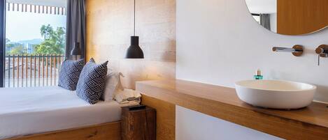 Une chambre avec un grand lit, une table de chevet, un miroir rond et une salle de bain avec un lavabo blanc.