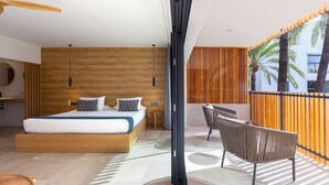 Un dormitorio moderno con una cama grande, cabecera de madera, mesita de noche pequeña y vistas a palmeras desde un balcón con baranda de madera.