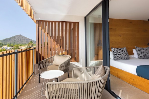 A balcony with wicker furniture, a bed with patterned pillows, and a view of a mountain. - Las Gaviotas Suites Hotel (Muro)