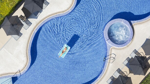 Vista aérea de una piscina con una fuente central, rodeada de sillas de descanso y sombrillas.