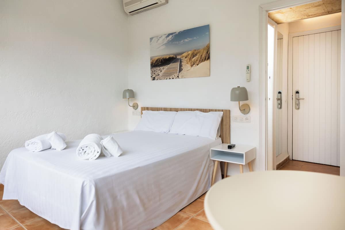 A bedroom with a bed, a bedside table, a wall-mounted lamp, a door, and a picture of a beach.