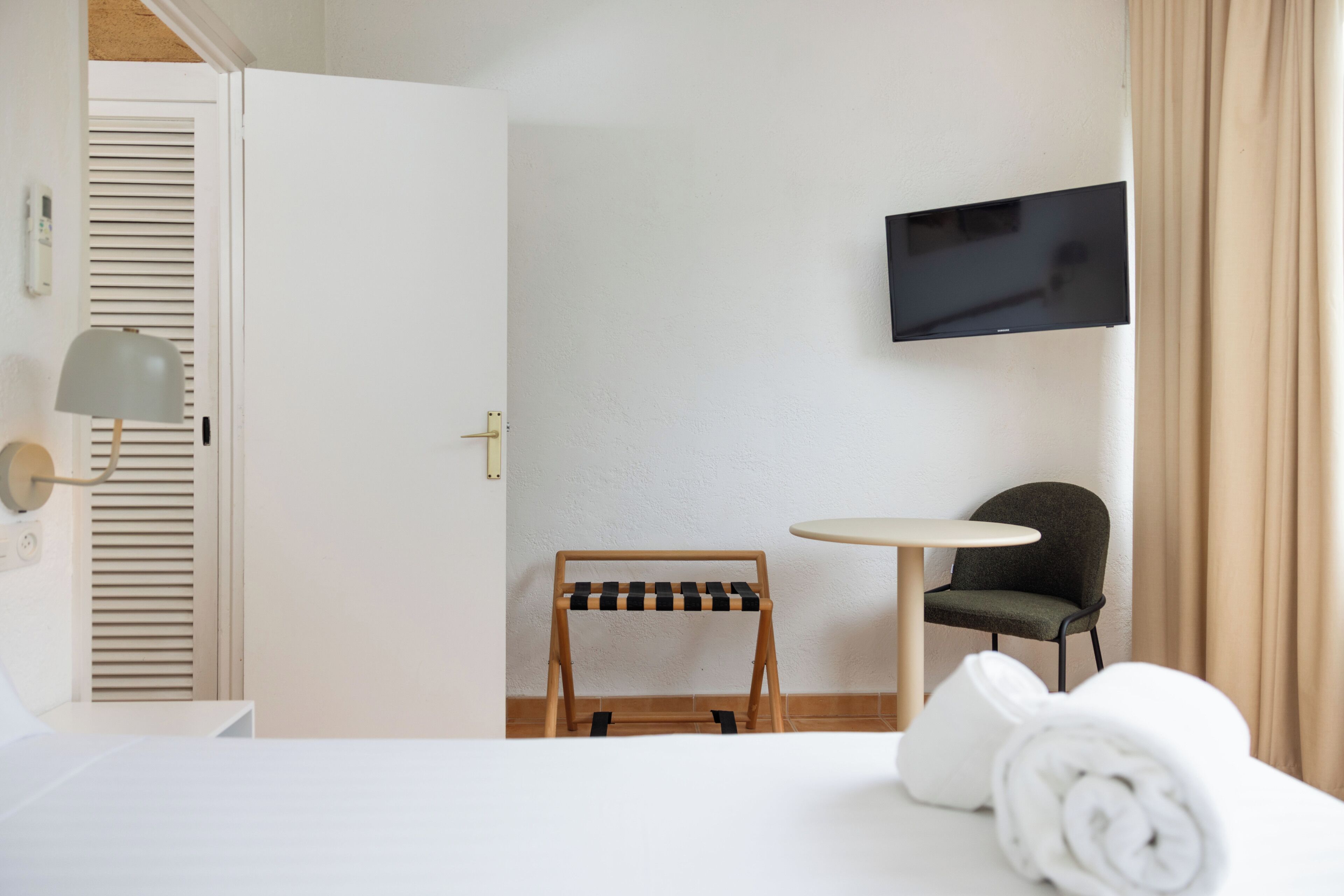 Une chambre d’hôtel avec un lit, un téléviseur fixé au mur, une petite table, une chaise et un porte-serviettes en bois.