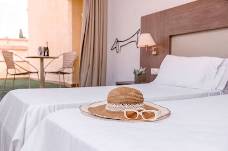 Una habitación de hotel con dos camas, una mesa con sillas, una lámpara y un sombrero con gafas de sol sobre una cama.. RVHotels Golf Costa Brava