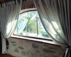 A window with white curtains, a stone wall, and a view of trees and greenery outside.