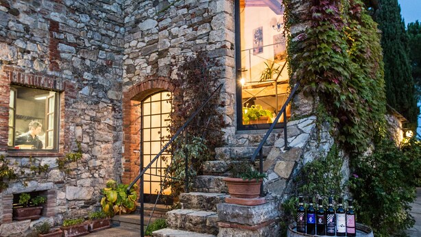 A stone building with a staircase leading to a lit entrance, potted plants, and a wooden barrel. - Borgo Livernano (Radda in Chianti)