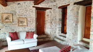 A rustic stone-walled room with a white sofa, red cushions, and a wooden door. - Borgo Livernano (Radda in Chianti)