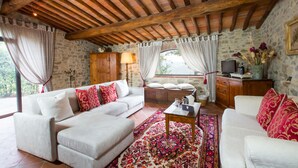 A cozy living room with a stone wall, wooden ceiling beams, and a large window with curtains. - Borgo Livernano (Radda in Chianti)