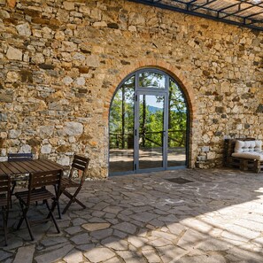 Un patio al aire libre con muros de piedra, una mesa de madera con sillas y una vista a través de un arco.