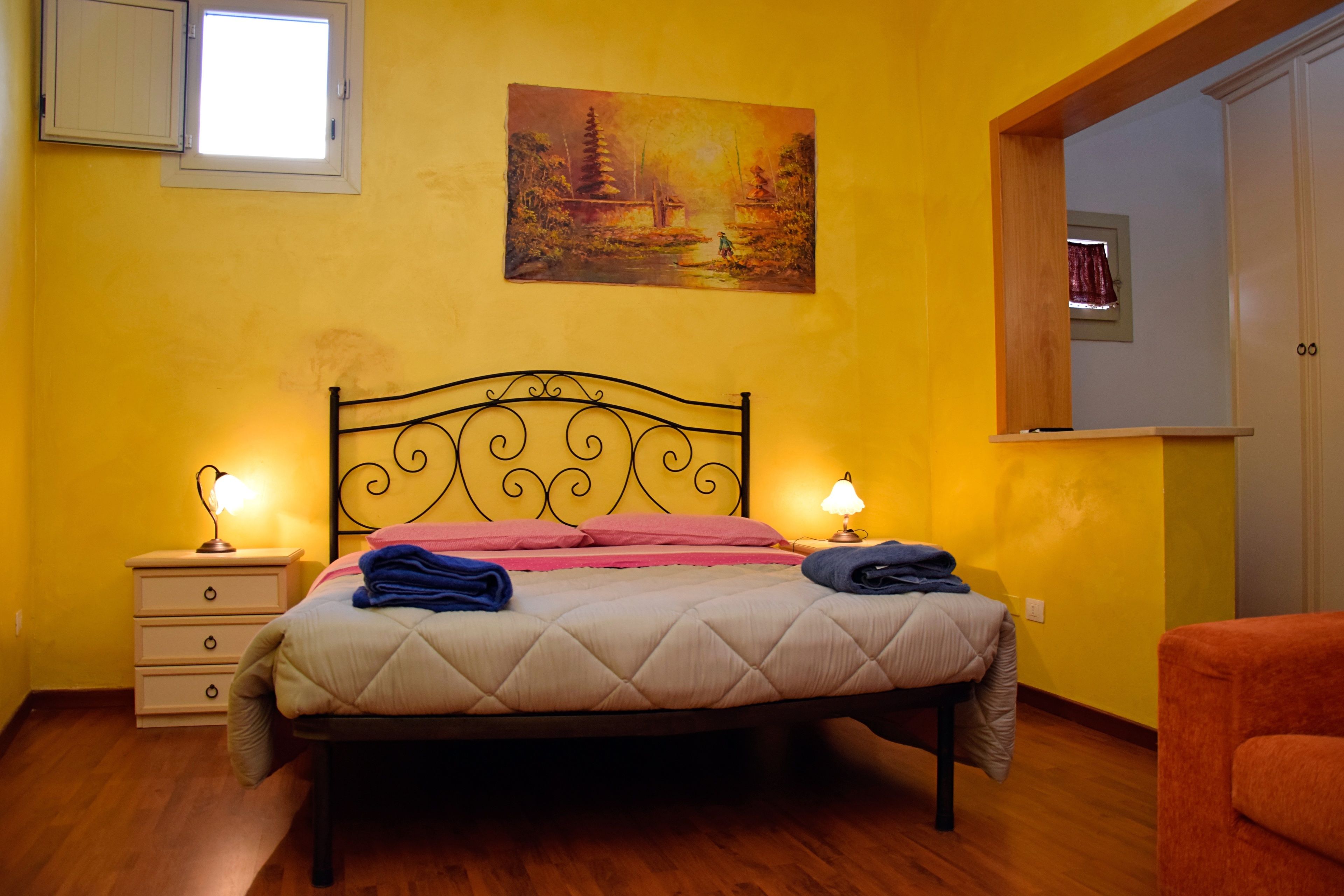 A bedroom with a bed, bedside lamps, a nightstand, a chair, a painting on the wall, and a window.