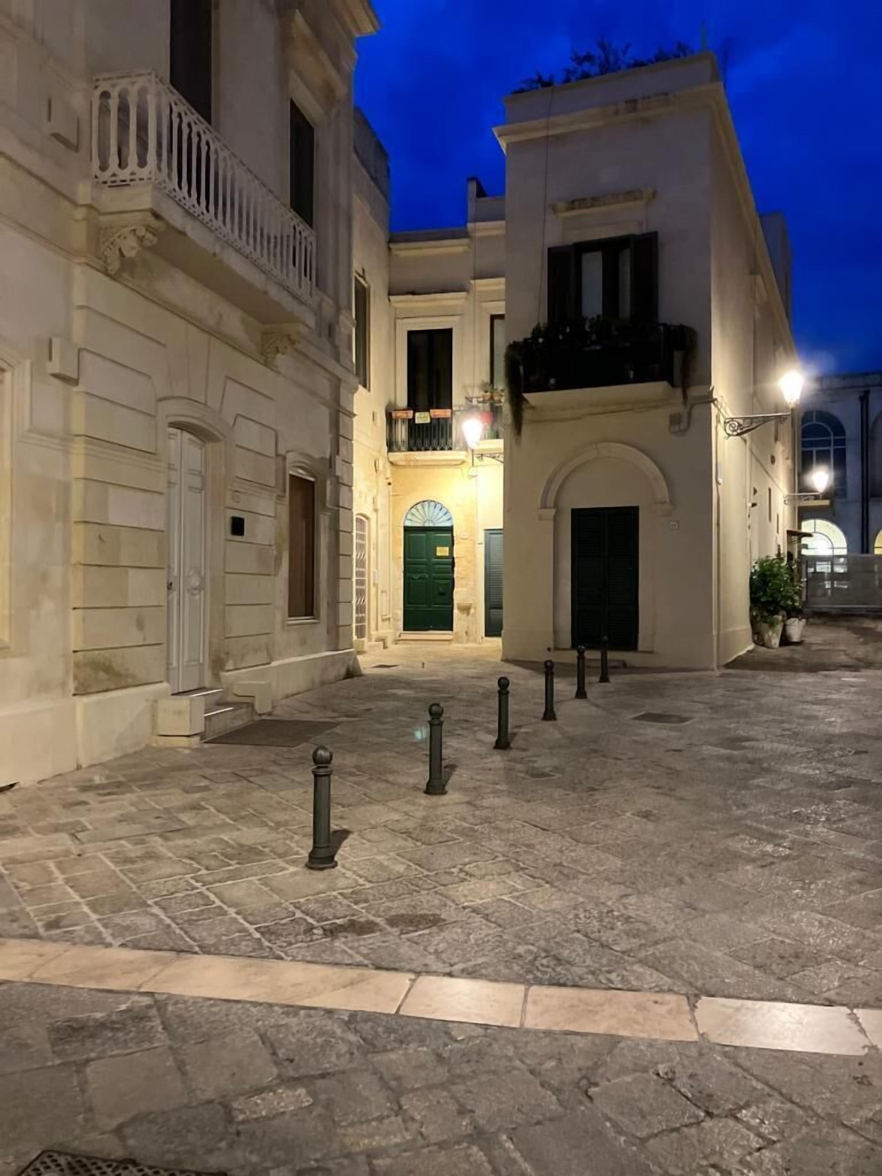A courtyard with stone flooring and bollards, surrounded by buildings with classical architecture and a green door.