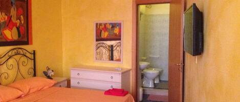 Un dormitorio con una cama, una mesita de noche, un cuadro, un televisor y una puerta abierta al baño.