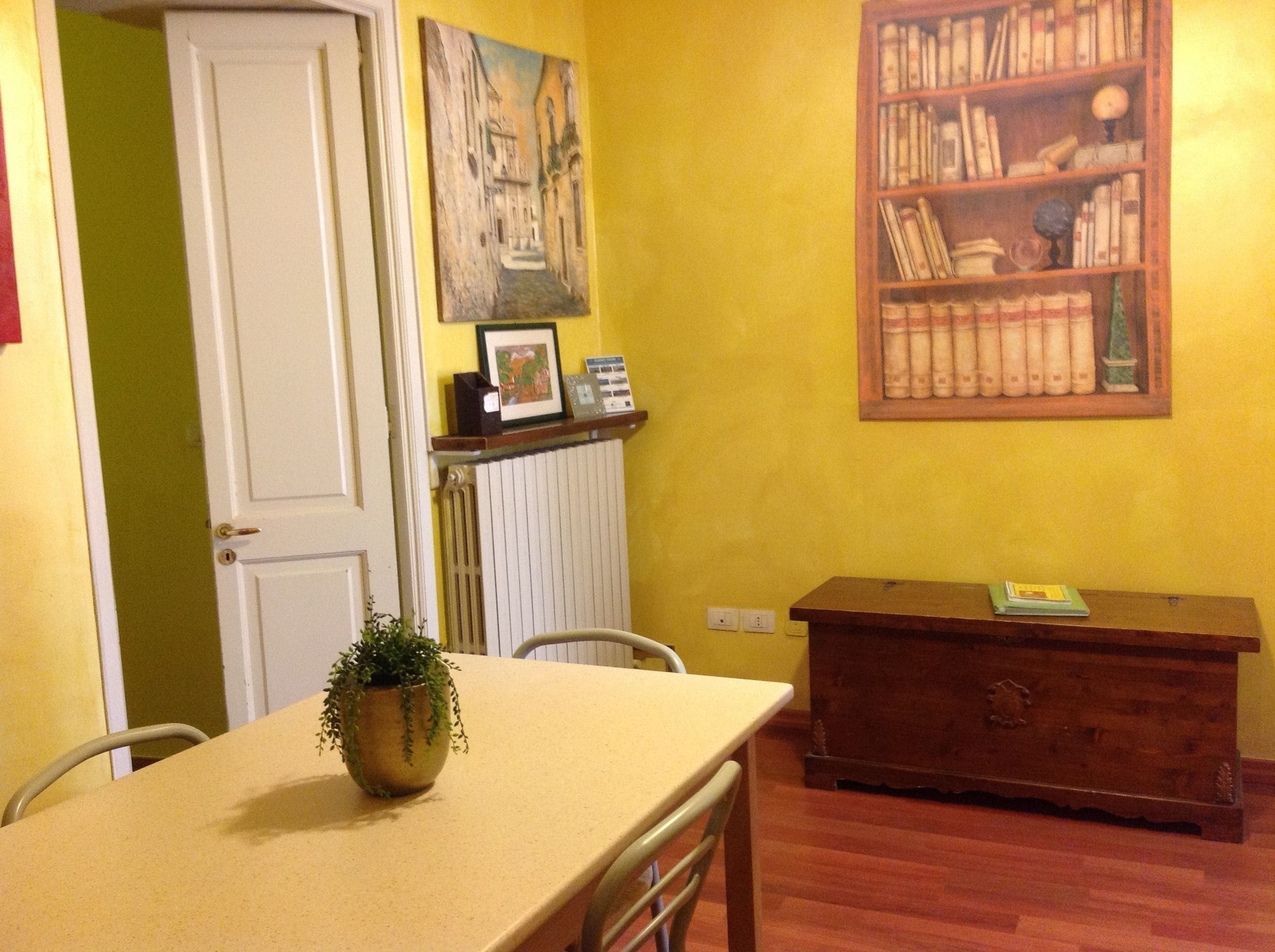 Een kamer met een houten tafel, een stoel, een kamerplant, een boekenkast en een radiator.