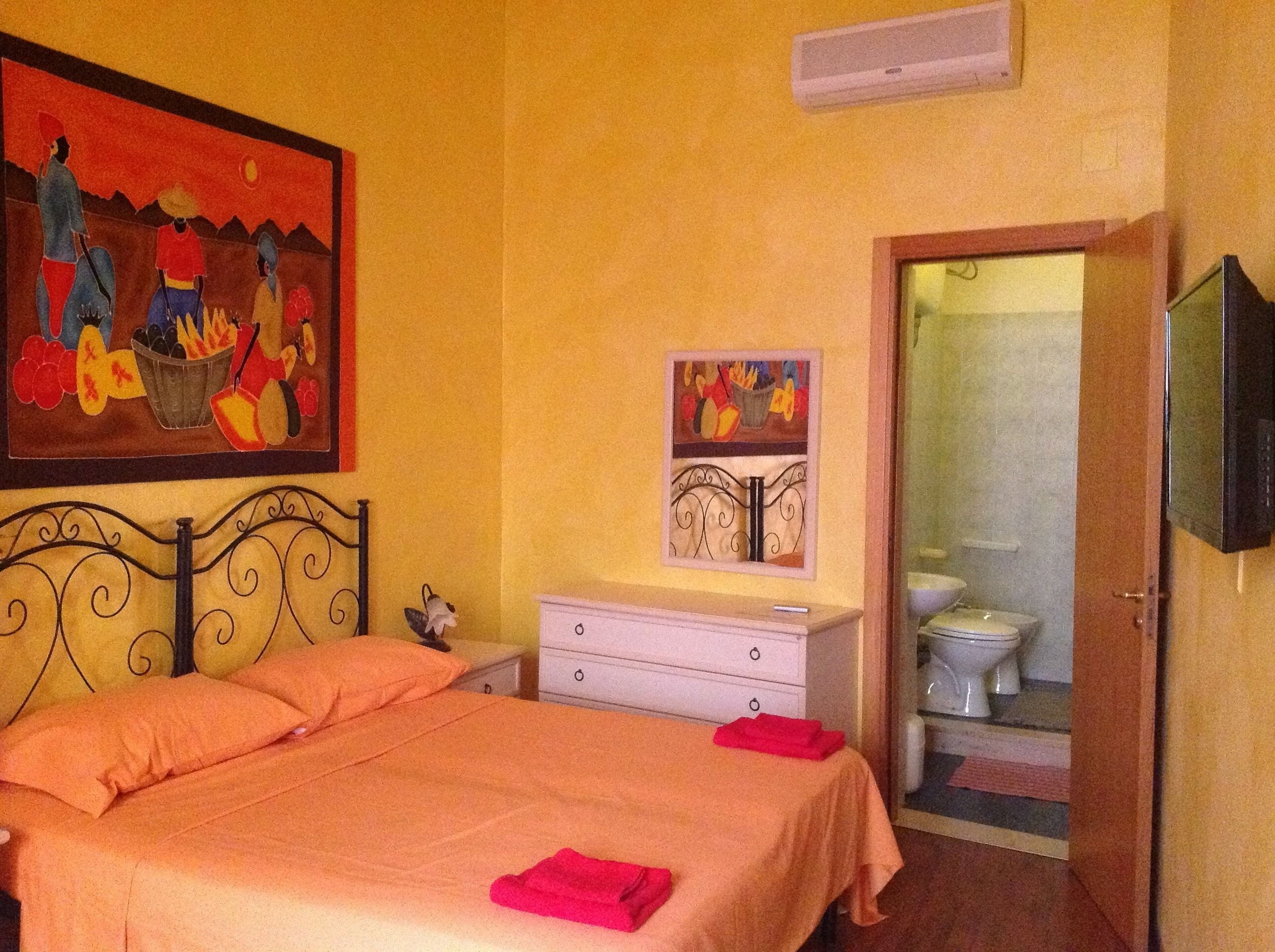A bedroom with a bed, a nightstand, a painting, a television, and a bathroom visible through an open door.