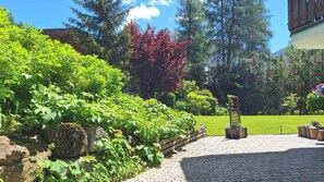 A paved patio area with a wooden fence, a stone wall, and a variety of green plants and trees. - Landhaus Charlotte (Seefeld in Tirol)