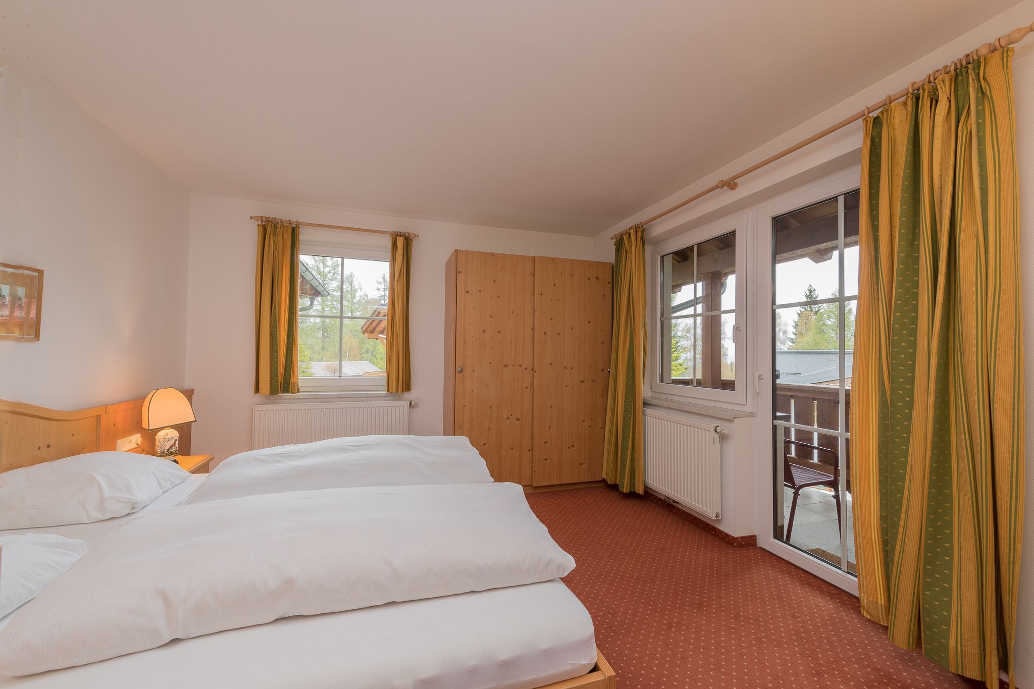 A hotel room with two beds, a wooden wardrobe, a window with curtains, and a radiator.