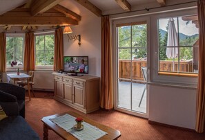 A cozy living room with a wooden ceiling, a television, a dining table with chairs, and large windows with curtains.