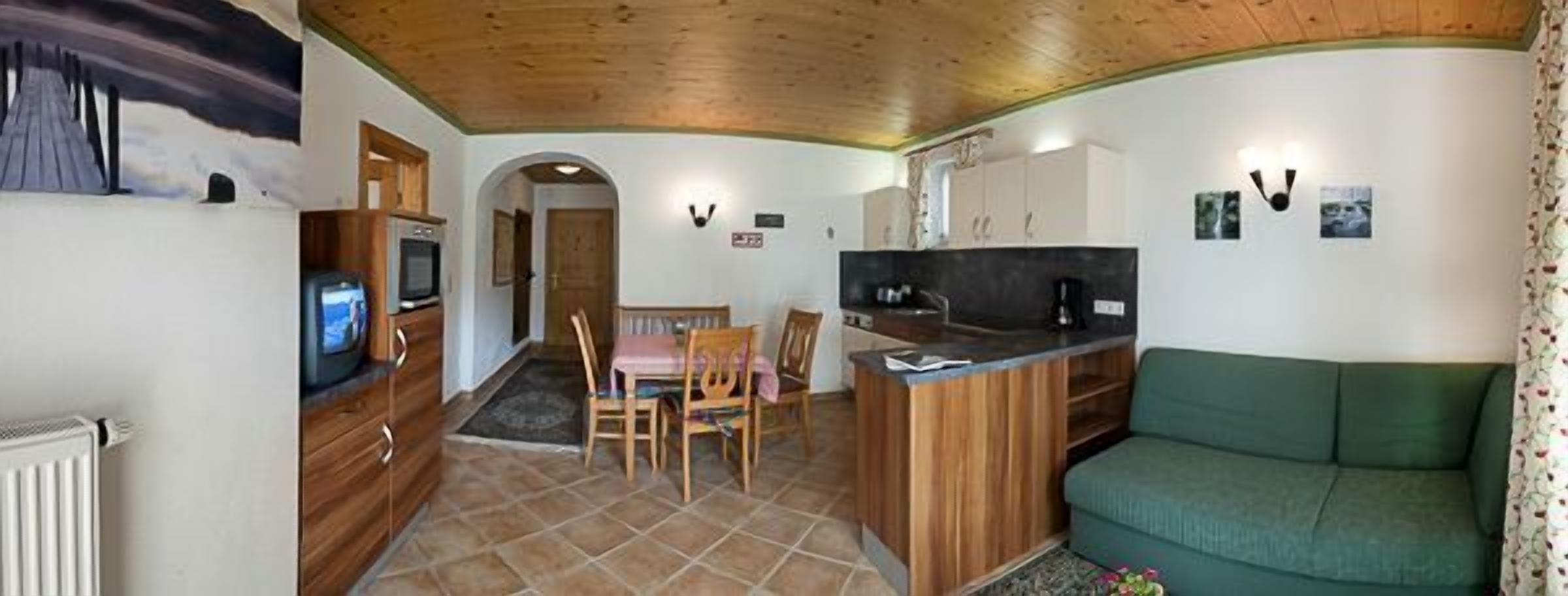 Un salon chaleureux avec un plafond en bois, un canapé vert, une table à manger avec des chaises, un coin cuisine doté d’un plan de travail noir et un téléviseur posé sur un meuble en bois.