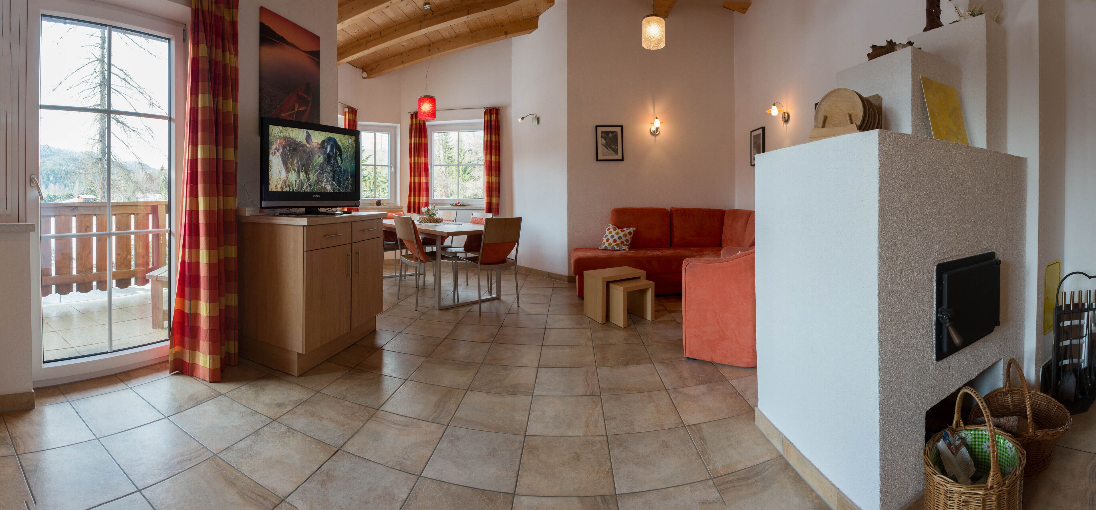 A spacious living area with a kitchen, dining table, and a red sofa. A television is mounted on the wall, and there are wicker baskets on the floor.