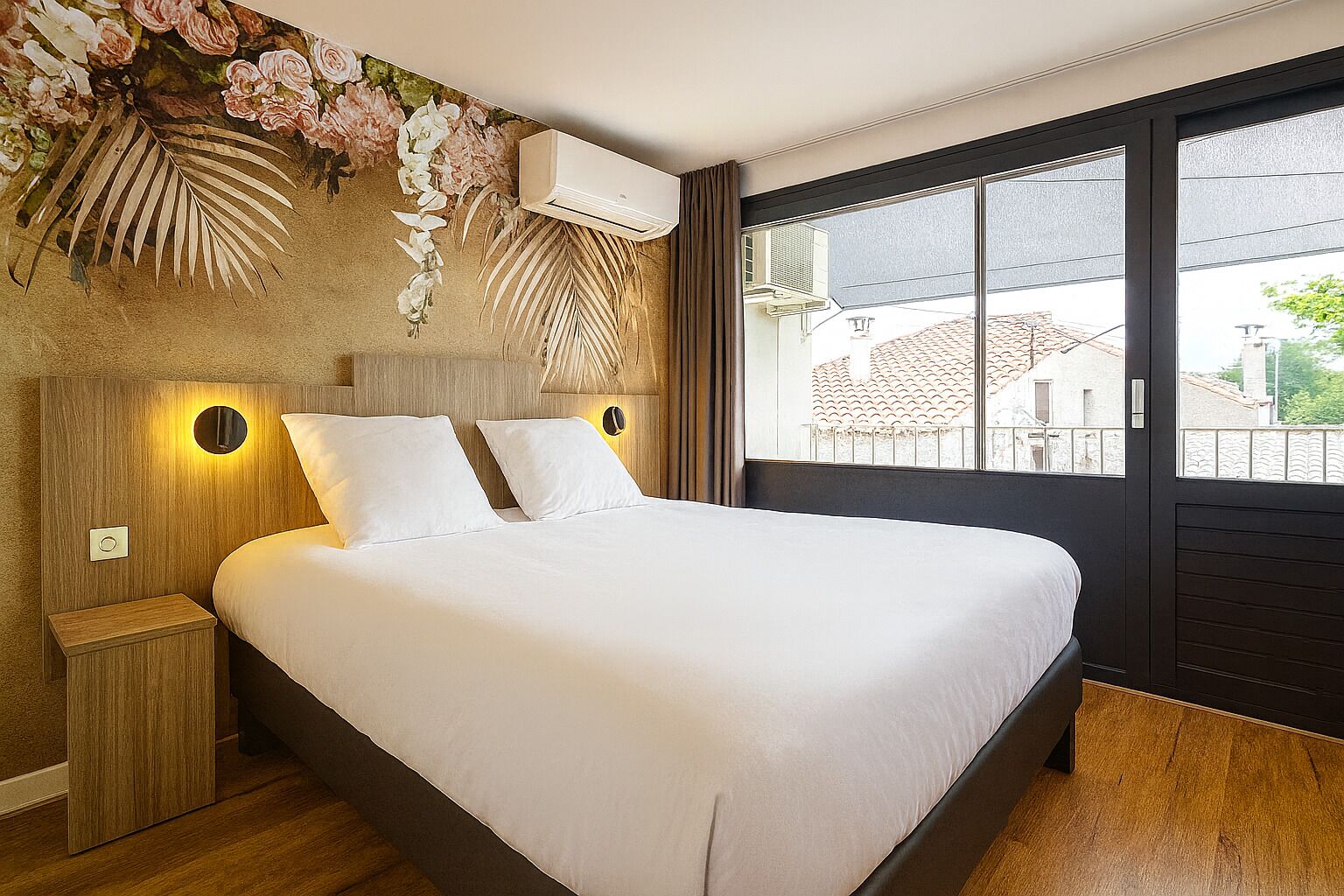 A bedroom with a large bed, wooden headboard, wall-mounted air conditioning, and a view of buildings through a large window.