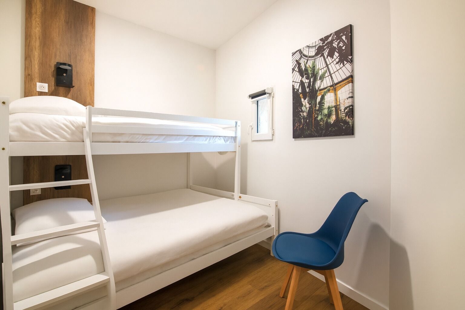 Dormitorio con literas, cabecera de madera, una silla azul y ventana con vista a un edificio.