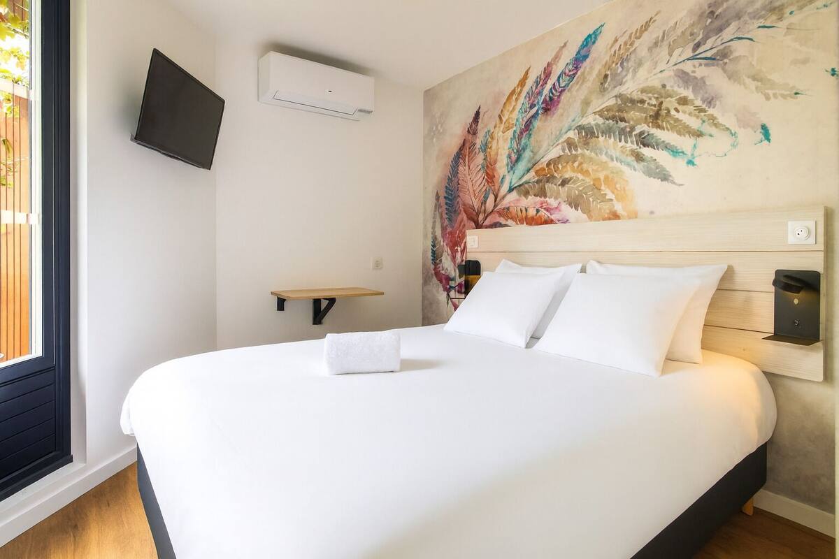 A hotel room with a large bed, a mounted TV, a wall-mounted air conditioner, and a wooden shelf.