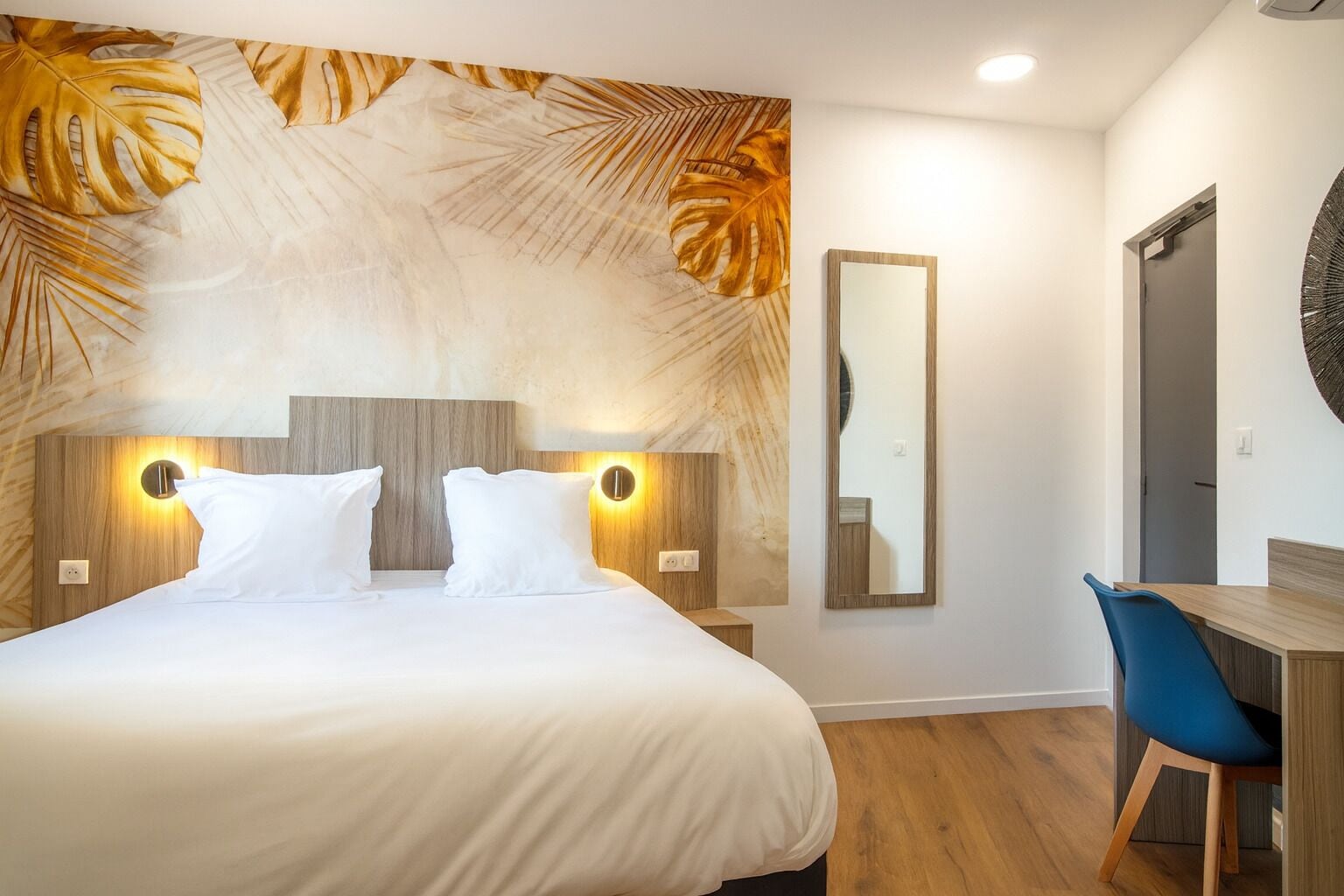 A modern hotel room with a large bed, two bedside tables with wall-mounted lamps, a desk with a chair, and a large mirror on the wall.