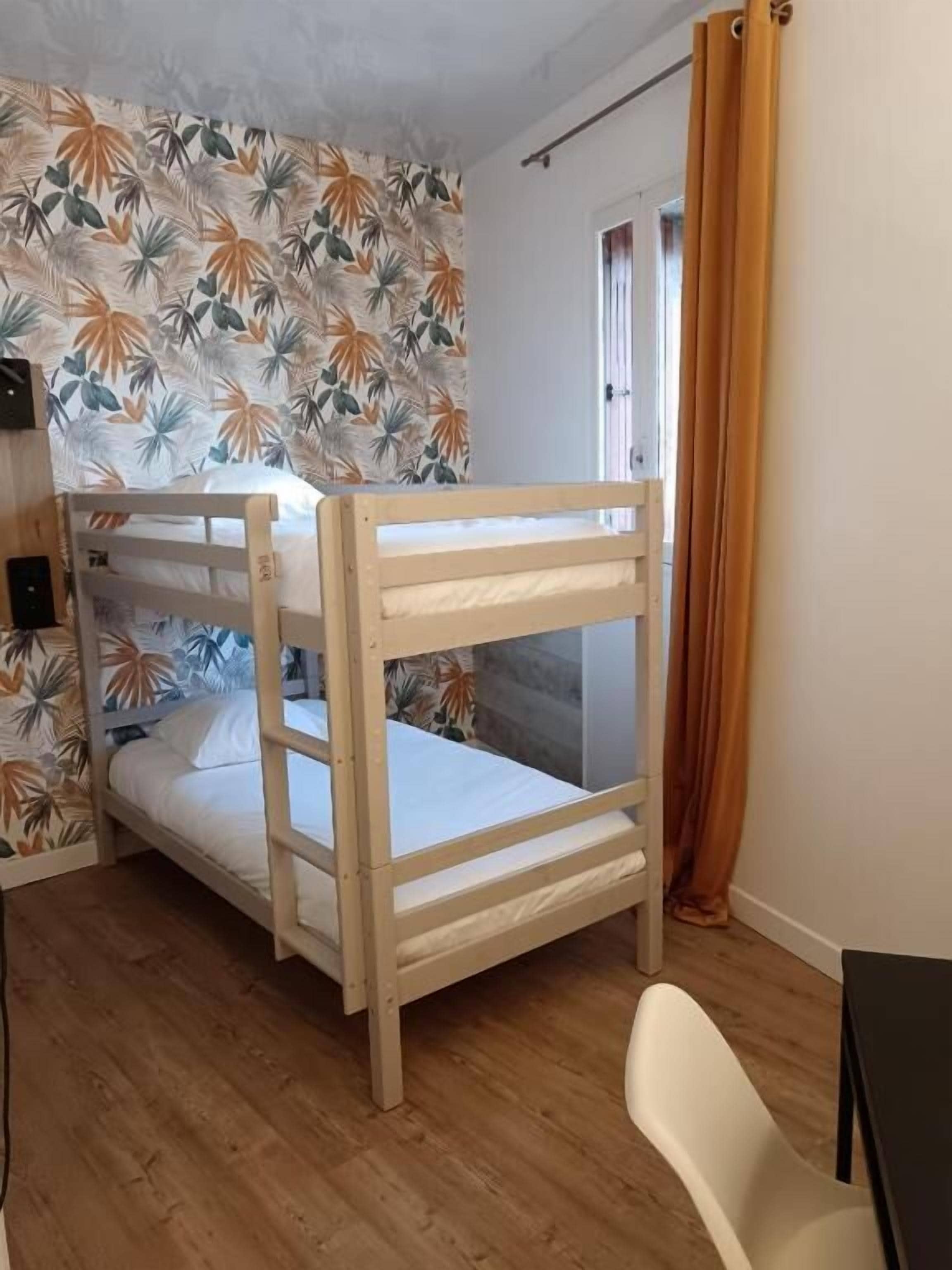 Een kamer met twee stapelbedden, een houten vloer, een witte muur met een tropisch bladpatroon en een deur voorzien van een gordijn.