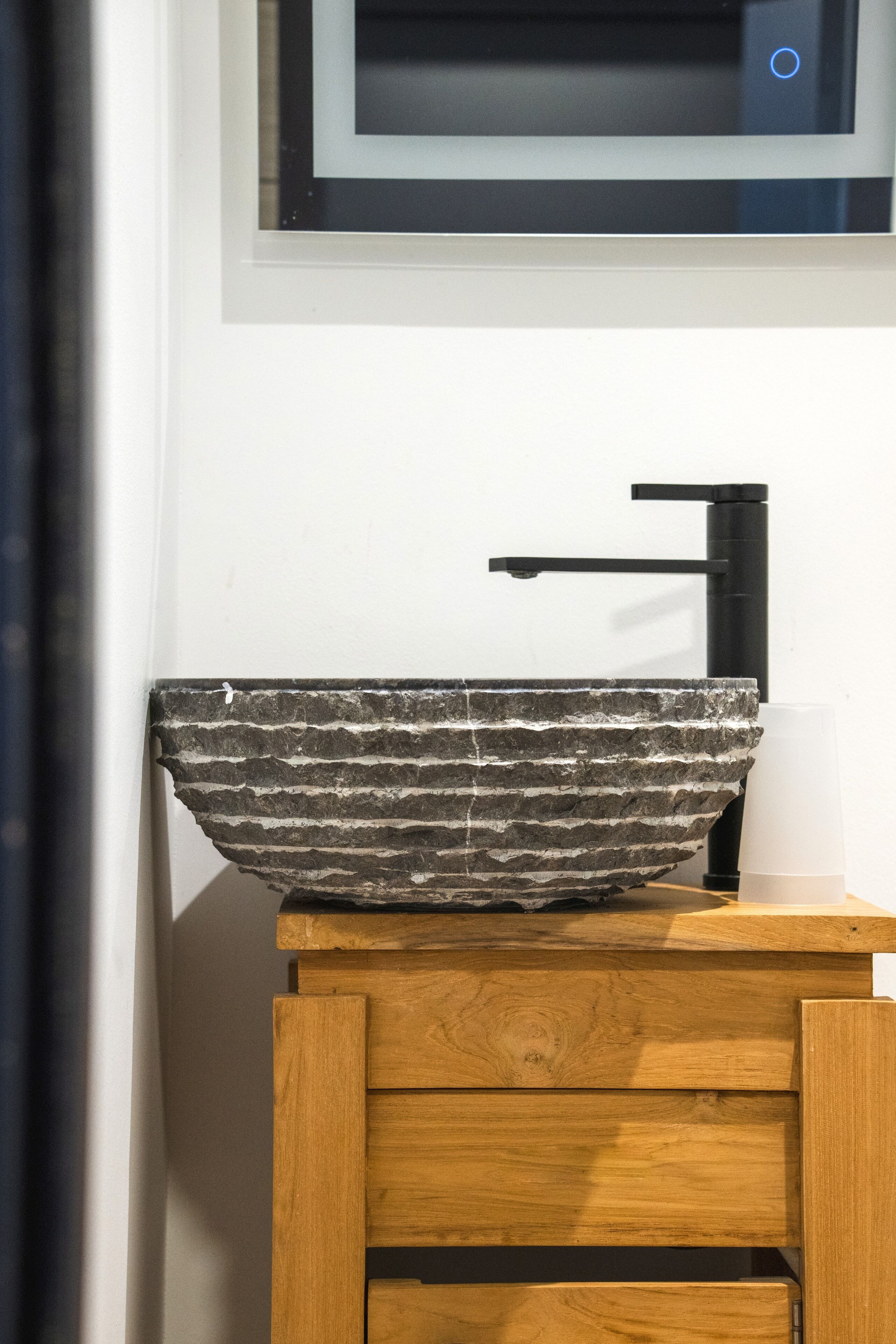 A stone vessel sink on a wooden pedestal with a black faucet, mounted on a white wall above a window.
