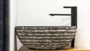 A stone vessel sink on a wooden pedestal with a black faucet, mounted on a white wall above a window.