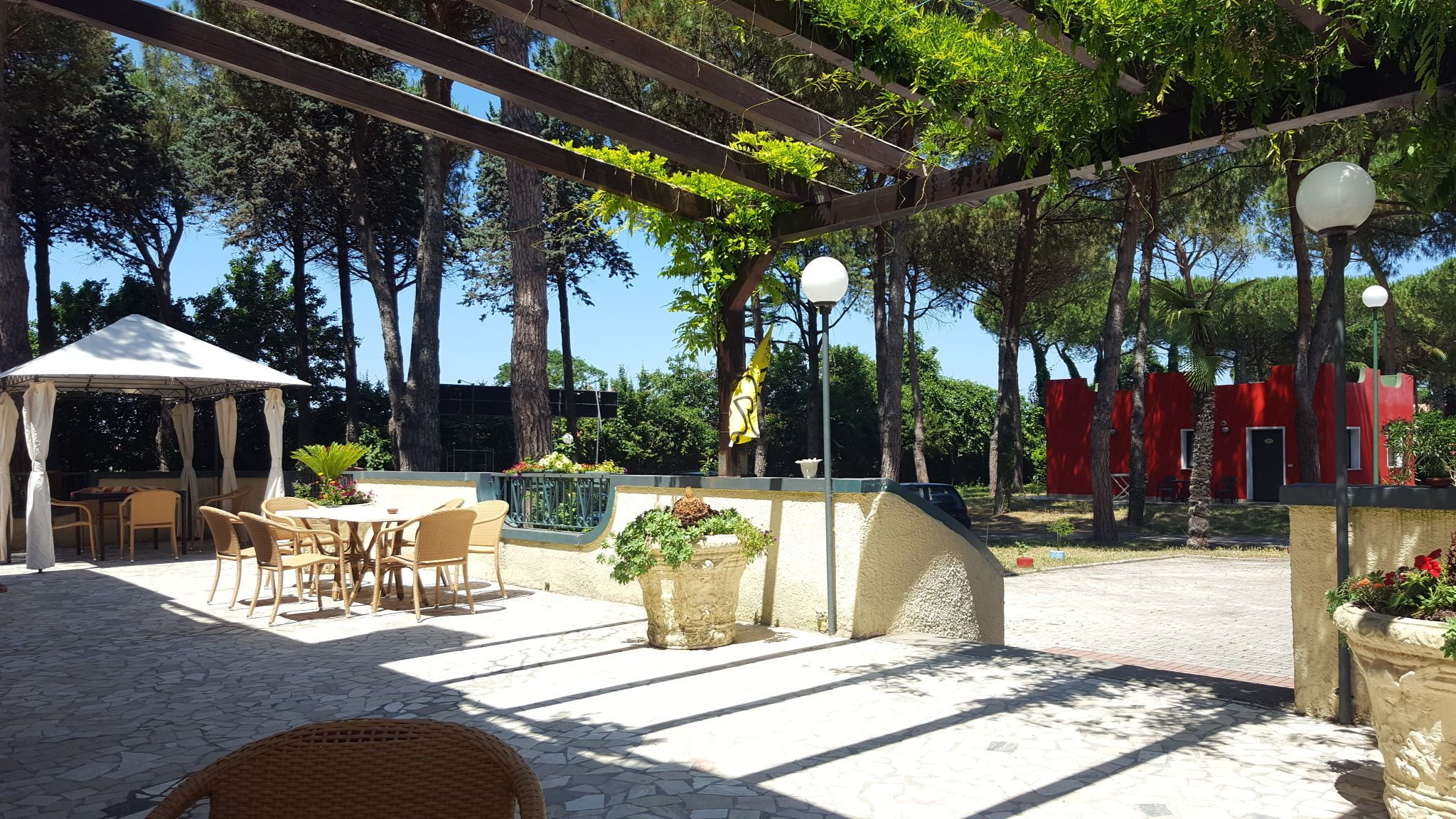 Un patio exterior con zona de estar cubierta, suelo de piedra y una pérgola con plantas colgantes.