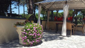 Zona d'estar a l'aire lliure coberta, amb paviment de pedra, una gran jardiniera amb flors roses i una pérgola amb teulada blanca.