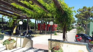 Ein überdachter Außenbereich mit einer Pergola, Kübelpflanzen und einem roten Gebäude im Hintergrund.