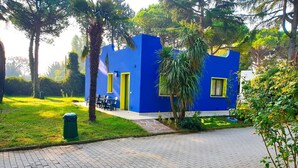 Edifici blau amb portes i finestres grogues, envoltat de vegetació i un camí pavimentat.