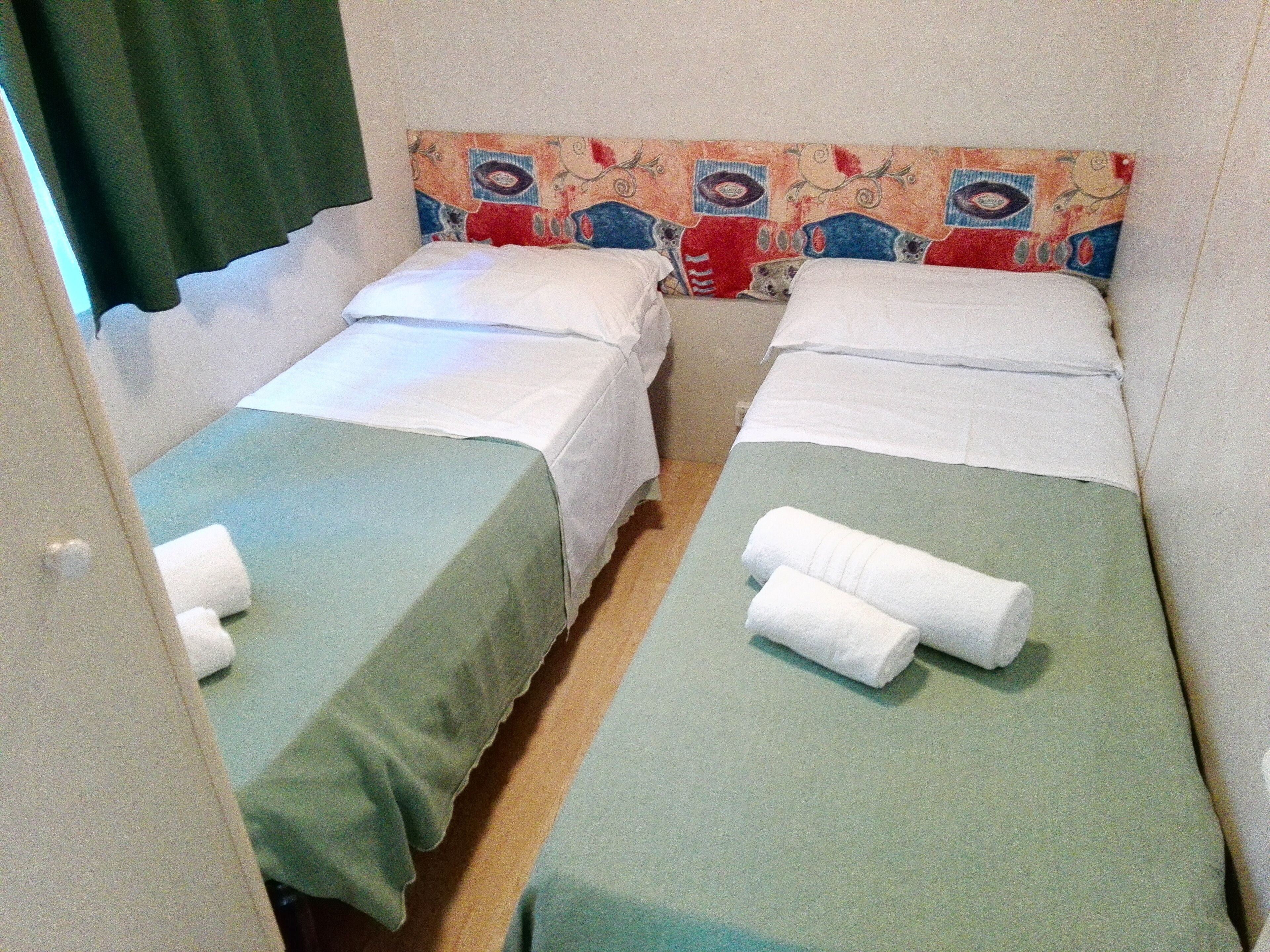 Ein Zimmer mit zwei Einzelbetten, jeweils mit grüner Bettdecke und weißen Laken, und einem farbenfrohen Kopfteil.