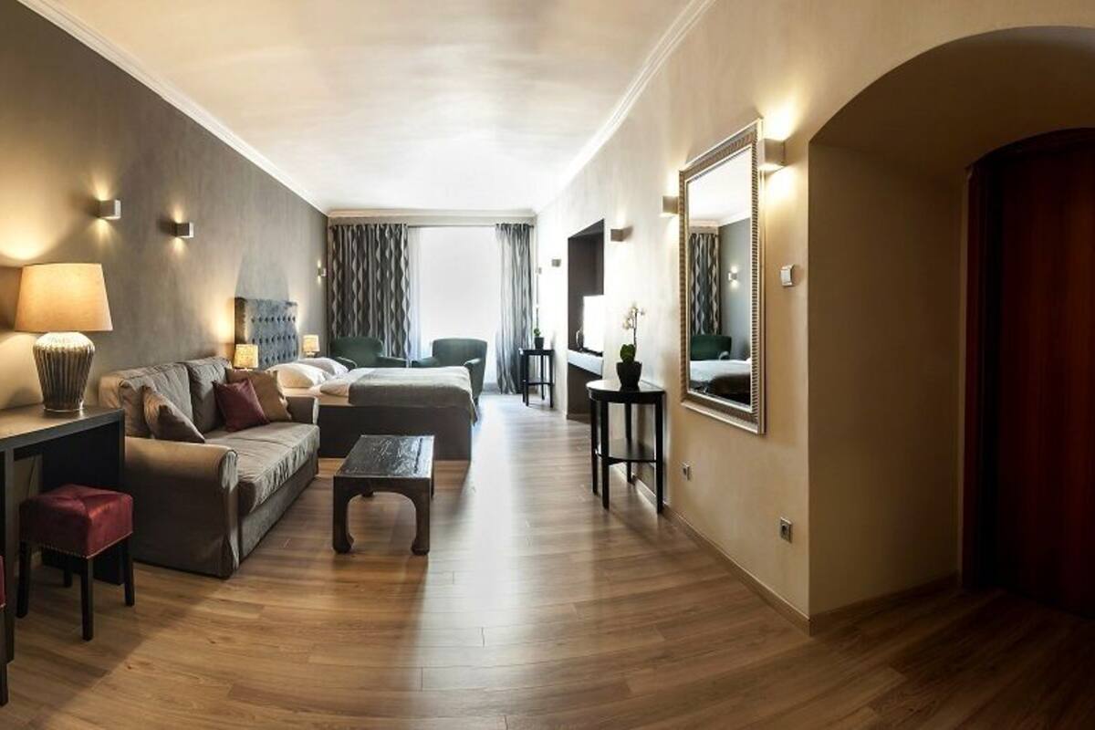 A modern hotel room with a sofa, two armchairs, a coffee table, a desk with a lamp, and a mirror.
