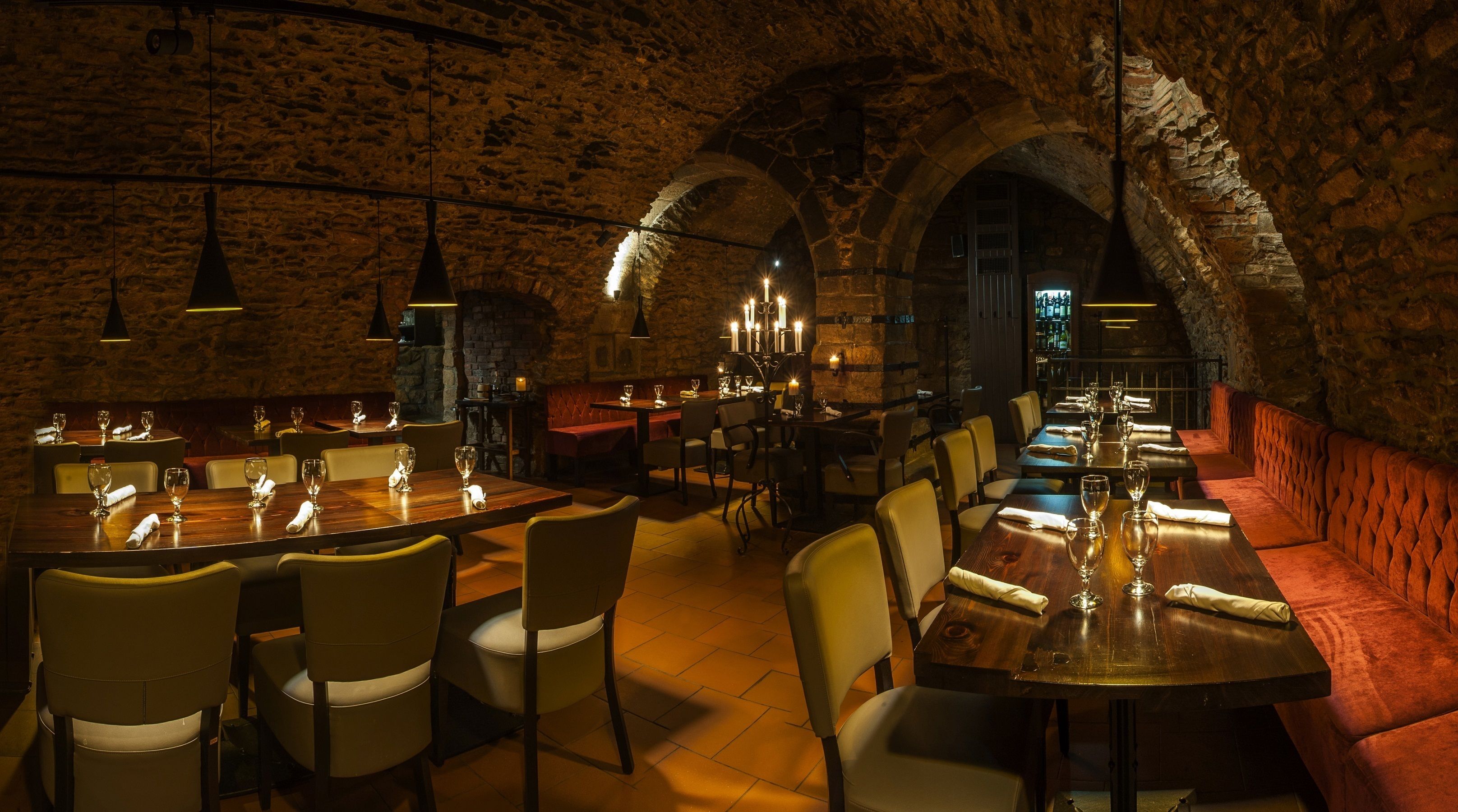 A dimly lit restaurant with stone walls and arched ceilings, featuring wooden tables and cushioned seating.