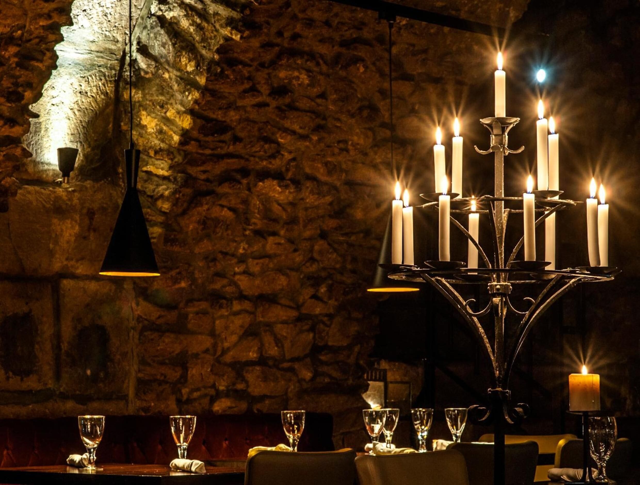 A candlelit table with multiple candles and glasses, set against a stone wall.