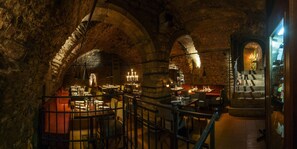 Un restaurant souterrain aux arches de pierre, où les tables sont dressées pour le dîner et où de magnifiques lustres illuminent la salle à la lueur des bougies.
