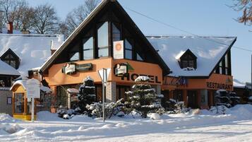 A hotel with a restaurant, featuring a large sign with a sun and snowflake logo, surrounded by snow-covered trees and a clear sky.