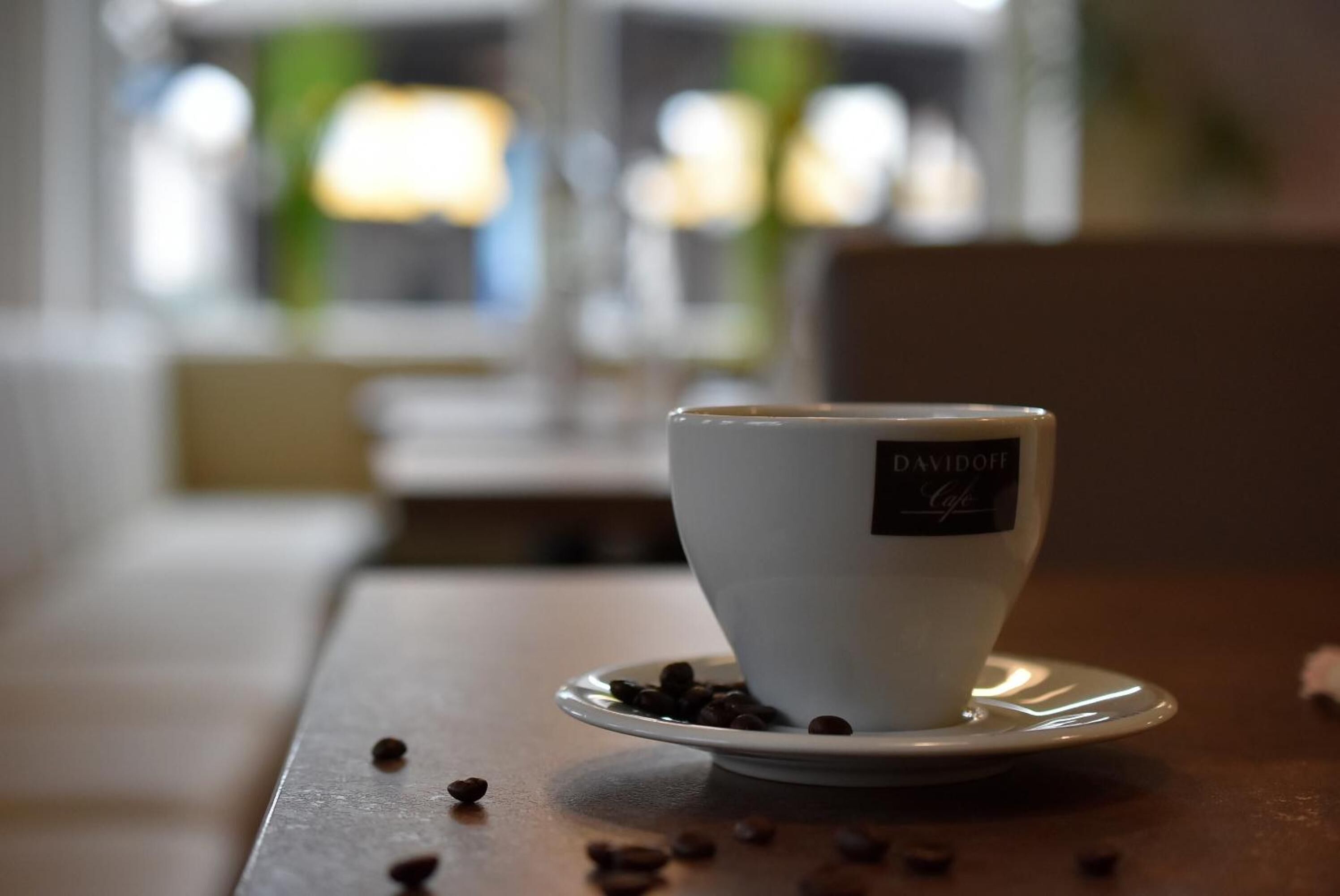 Una taza blanca con etiqueta sobre un platito, colocada sobre una mesa con granos de café esparcidos.