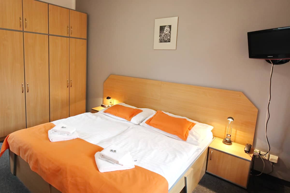 A hotel room with a bed, wooden wardrobe, bedside table, lamp, and a TV mounted on the wall.