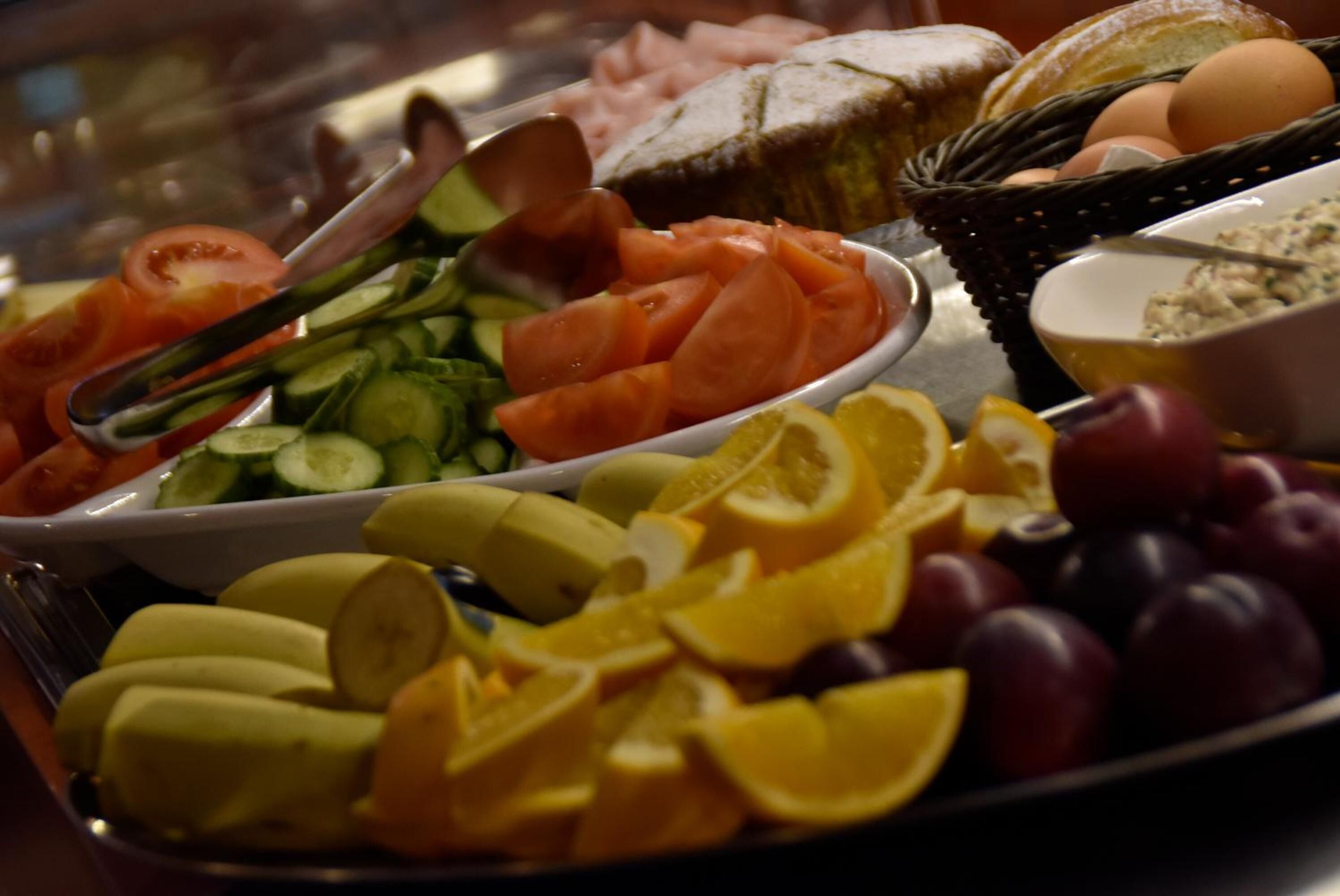 A buffet with various fruits and vegetables, including tomatoes, cucumbers, bananas, oranges, and grapes.
