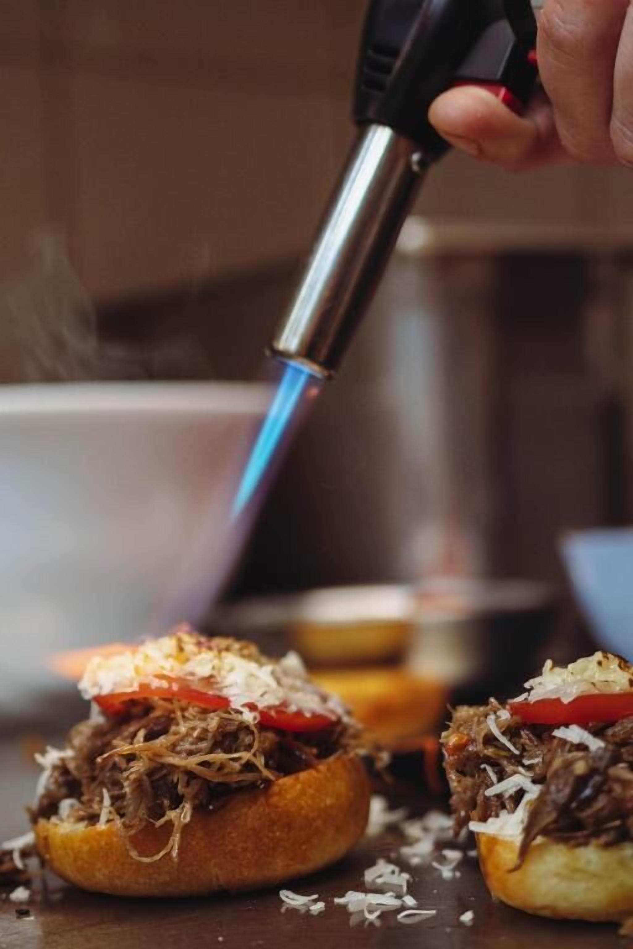 Gros plan d’une main tenant un chalumeau au-dessus d’un aliment, peut-être un sandwich ou une pâtisserie, avec des ingrédients visibles comme de la viande effilochée et une tranche de tomate.