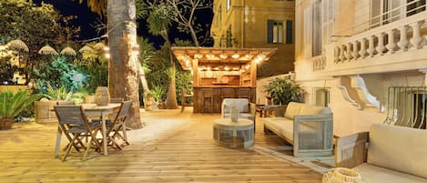A wooden deck with outdoor furniture, a covered seating area, and a building with balconies.