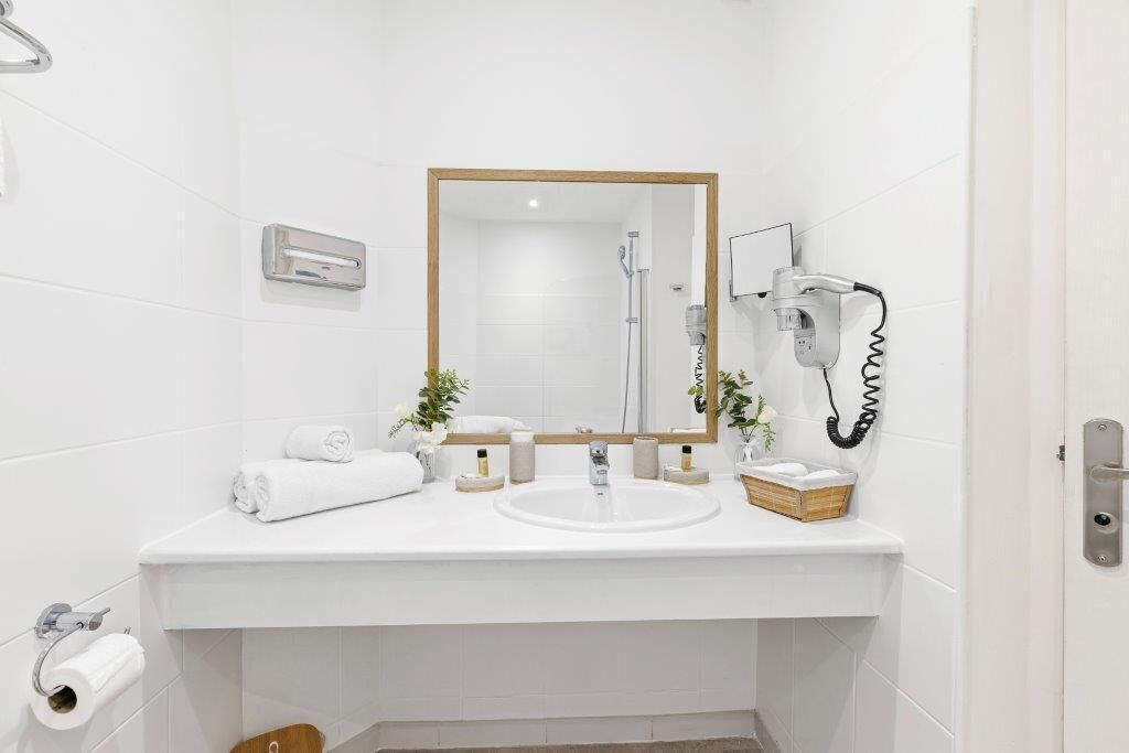 Un baño moderno con un lavamanos blanco, un espejo, un perchero para toallas y un secador de pelo en la pared.