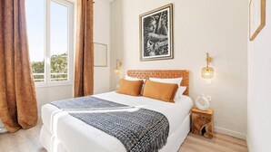 A bedroom with a bed, curtains, a framed picture, bedside lamps, and a small wooden nightstand. - Villa Claudia Hotel Cannes Centre - Parking (Cannes)