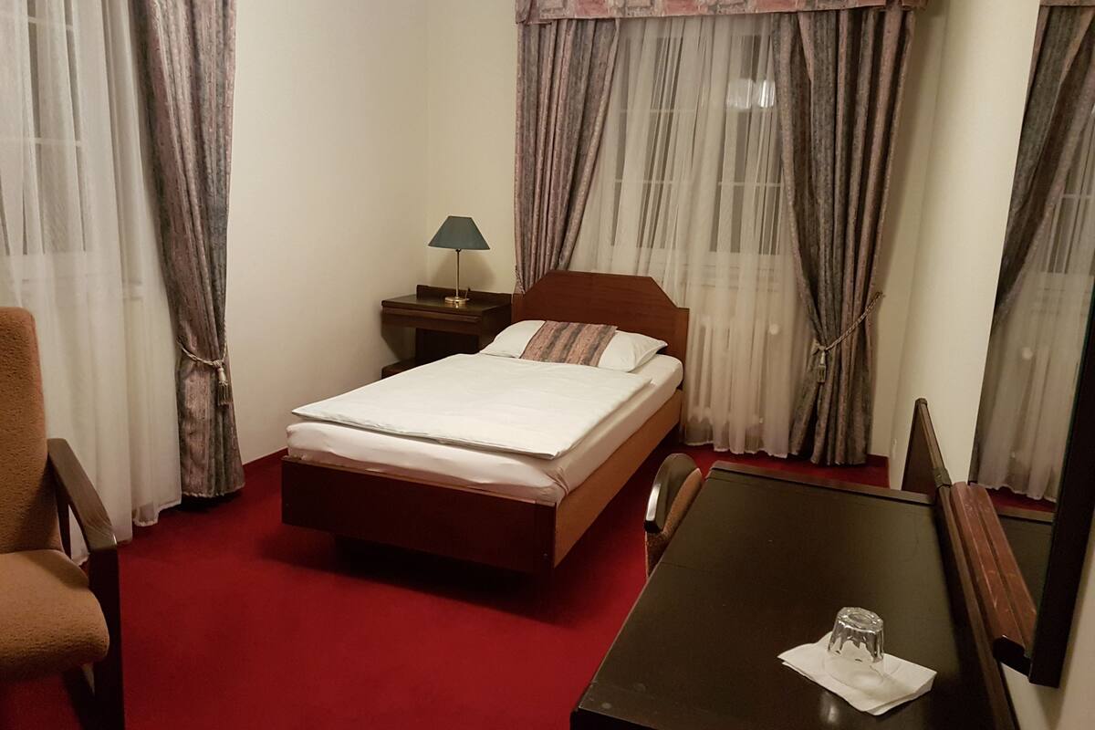 A hotel room with a bed, a nightstand with a lamp, a table with a glass and napkin, and a chair.