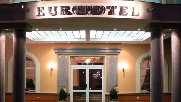 Der Eingang des Eurohotel mit Glastür, Säulen und einem grünen Teppichbereich.