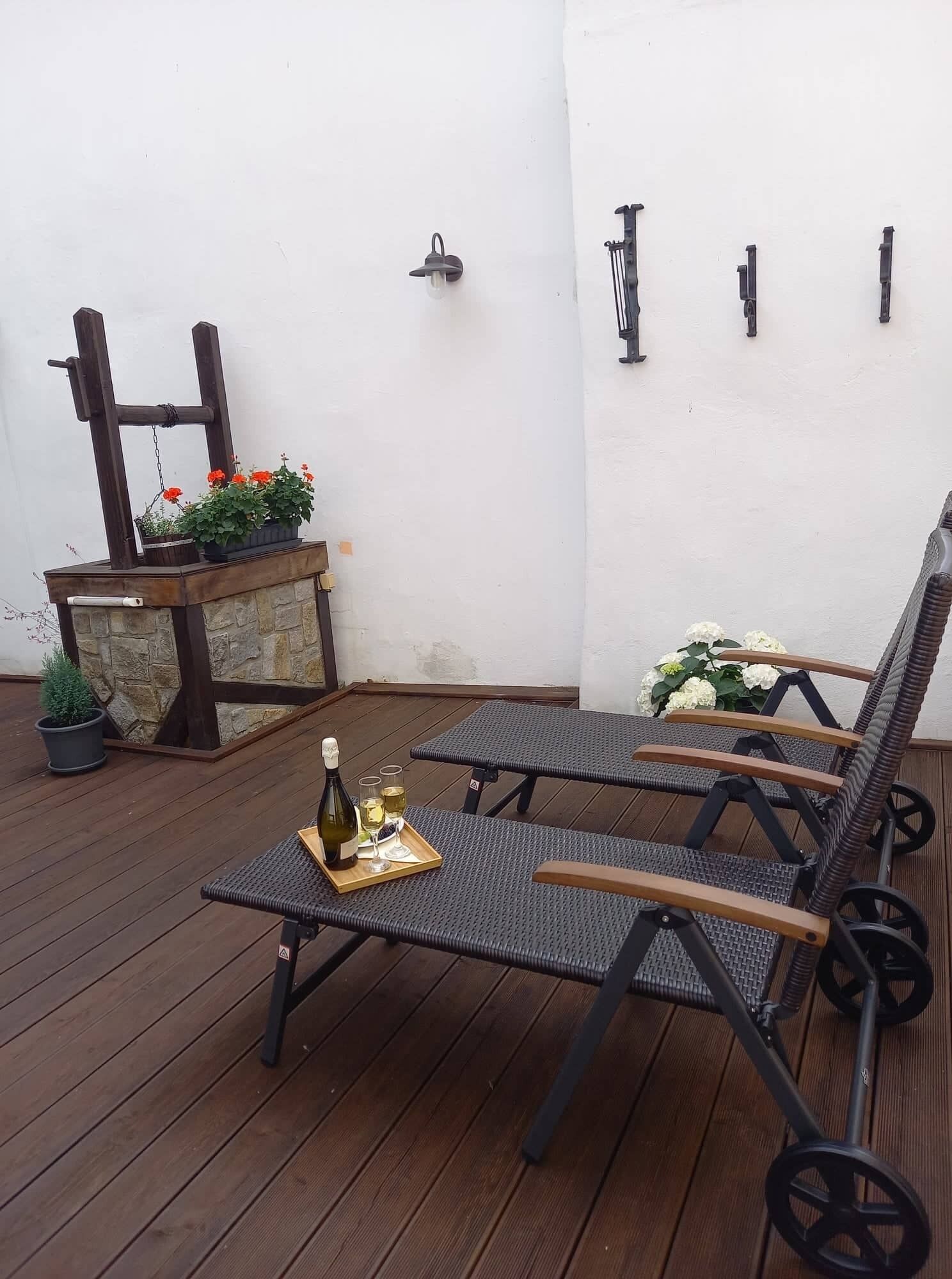 A wooden deck with two wicker lounge chairs, a small table with a bottle and glasses, and a stone planter with flowers.