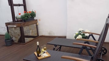 A wooden deck with two wicker lounge chairs, a small table with a bottle and glasses, and a stone planter with flowers.